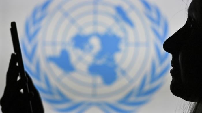 A woman looks at her cell phone in front of the United Nations logo displayed on a computer screen.