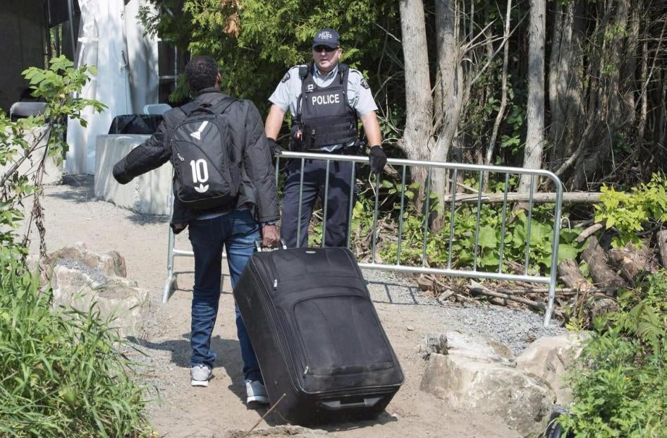 A migrant walks with his backpack and luggage and sees a Canadian police officer who holds onto a fence.