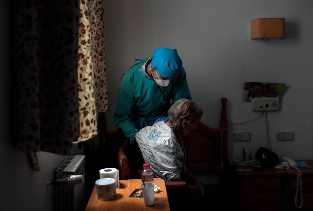 A doctor in a mask and scrubs checks on an elderly woman using a stethoscope