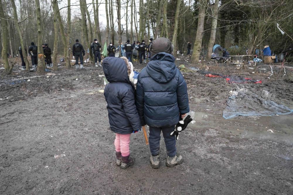 Two children wearing winter coats stand in a forest in front of a group of police officers