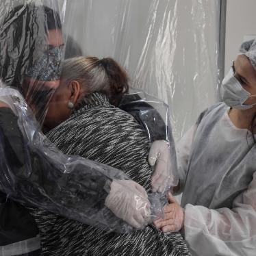 Two women in facemasks hug through a plastic curtain while a medical worker looks on