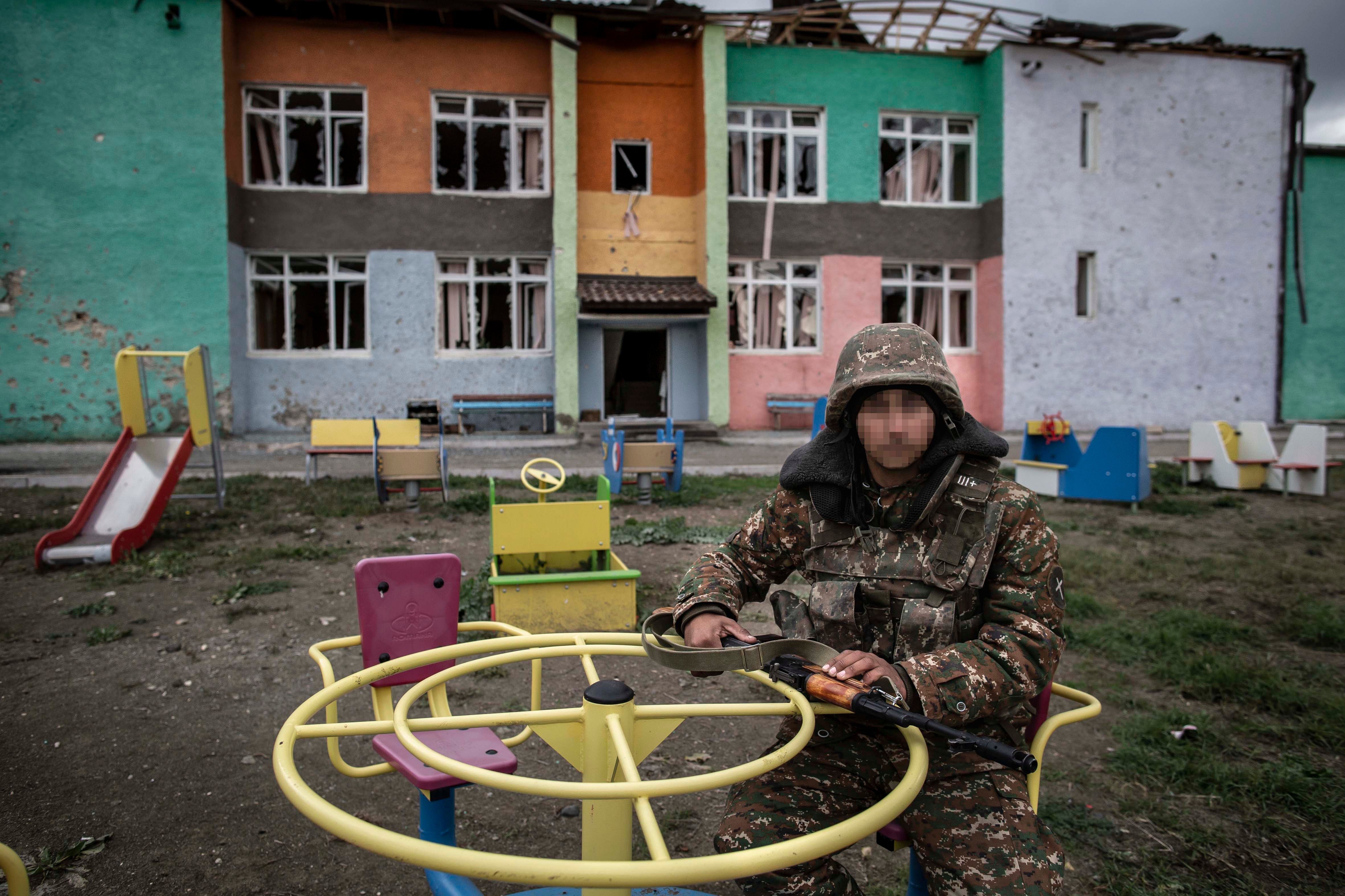 A soldier sits at playground equipment in front of a damaged school building