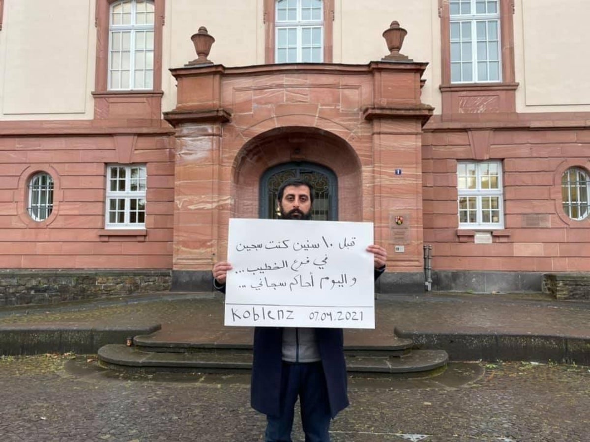 Amer Matar holds a poster with a message in Arabic that states, “I was jailed in al-Khatib branch, and today I am witnessing my jailor on trial.”