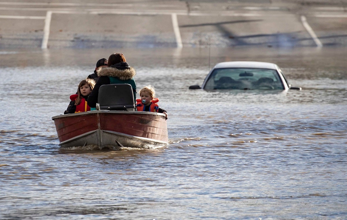 A woman and children in boat leaving behind car with floodwaters up to the roof in the background.