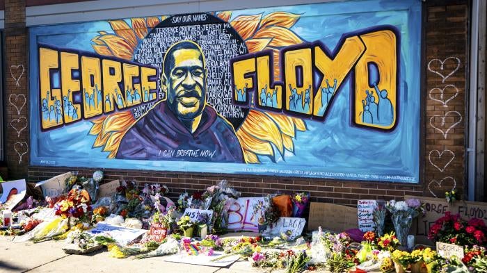 A mural dedicated to George Floyd and honoring the black lives matter movement, showing a depiction of George Floyd's face with his name and supporters of the movement against a blue painted background. Flowers and handwritten signs adorn the sidewalk beneath the mural.