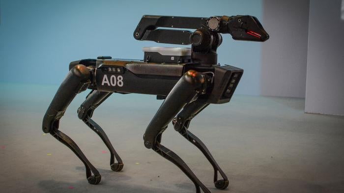 A black robot with four legs resembling a dog stands on a stage in front of a shite background.