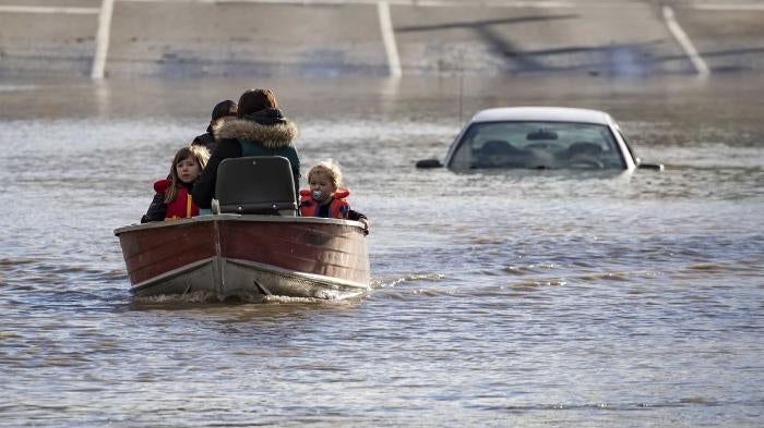 A woman and children in boat leaving behind car with floodwaters up to the roof in the background.