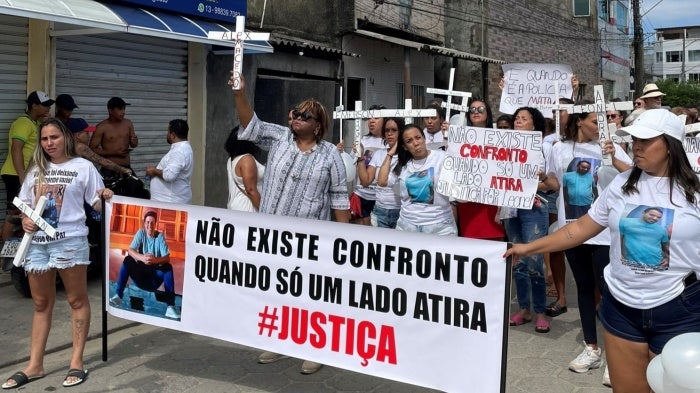  The image shows a group of people participating in a protest. The protesters hold banners and wear t-shirts with a person's face. One prominent banner read, in Portuguese, "There is no confrontation when only one side shoots" with the word "JUSTICE" emphasized below. 