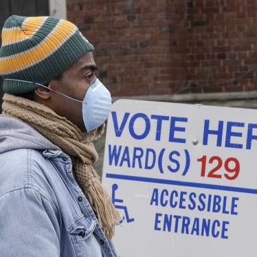 A man in a beanie and face mask stands in front of a sign that says "Vote Here"
